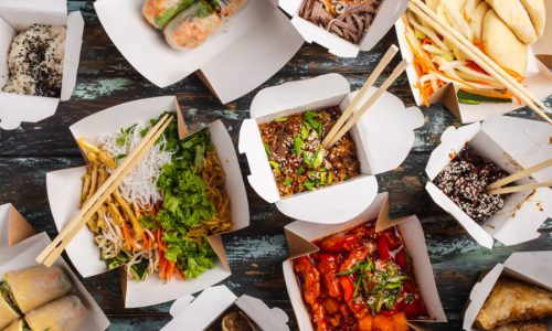 Assorted Chinese dishes in paper delivery boxes: sweet and sour chicken, dim sum, spring rolls, noodles, salad, rice, steamed buns, dips. Asian restaurant take away concept, top view